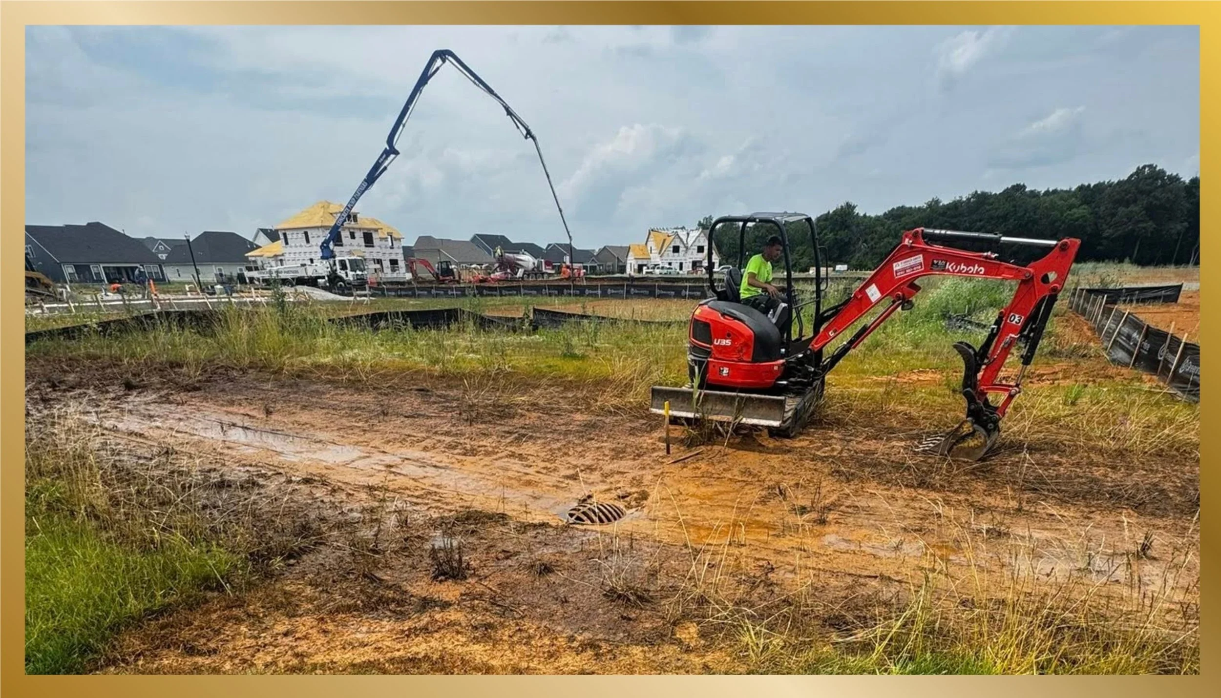 Construction site with a small red excavator and a worker operating it, houses in the background, and a truck with a crane lifting materials.