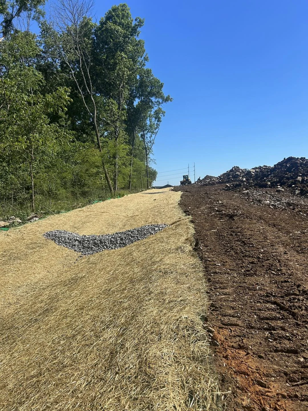 A construction or landscaping site showing a dirt pathway with a moisture barrier underneath. On the left, there are tall green trees and on the right, a pile of dirt and rocks. Equipment can be seen in the background under a clear blue sky.