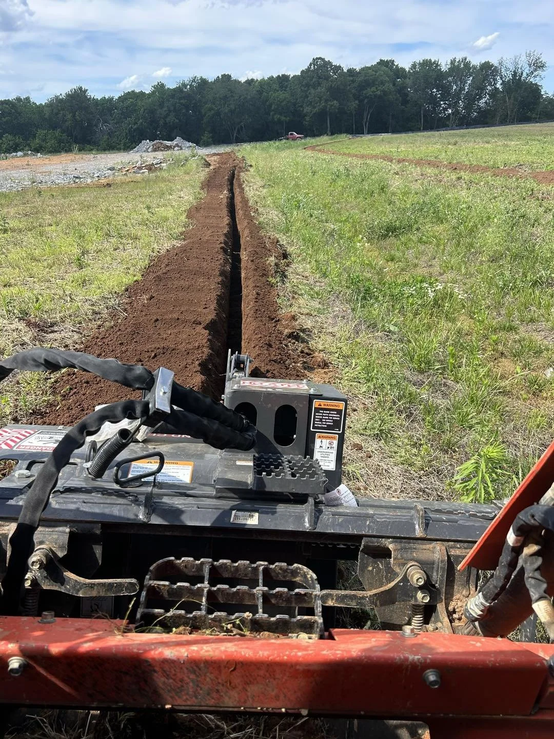 A tractor is making a trench in a grassy field. The field is surrounded by trees under a partly cloudy sky.