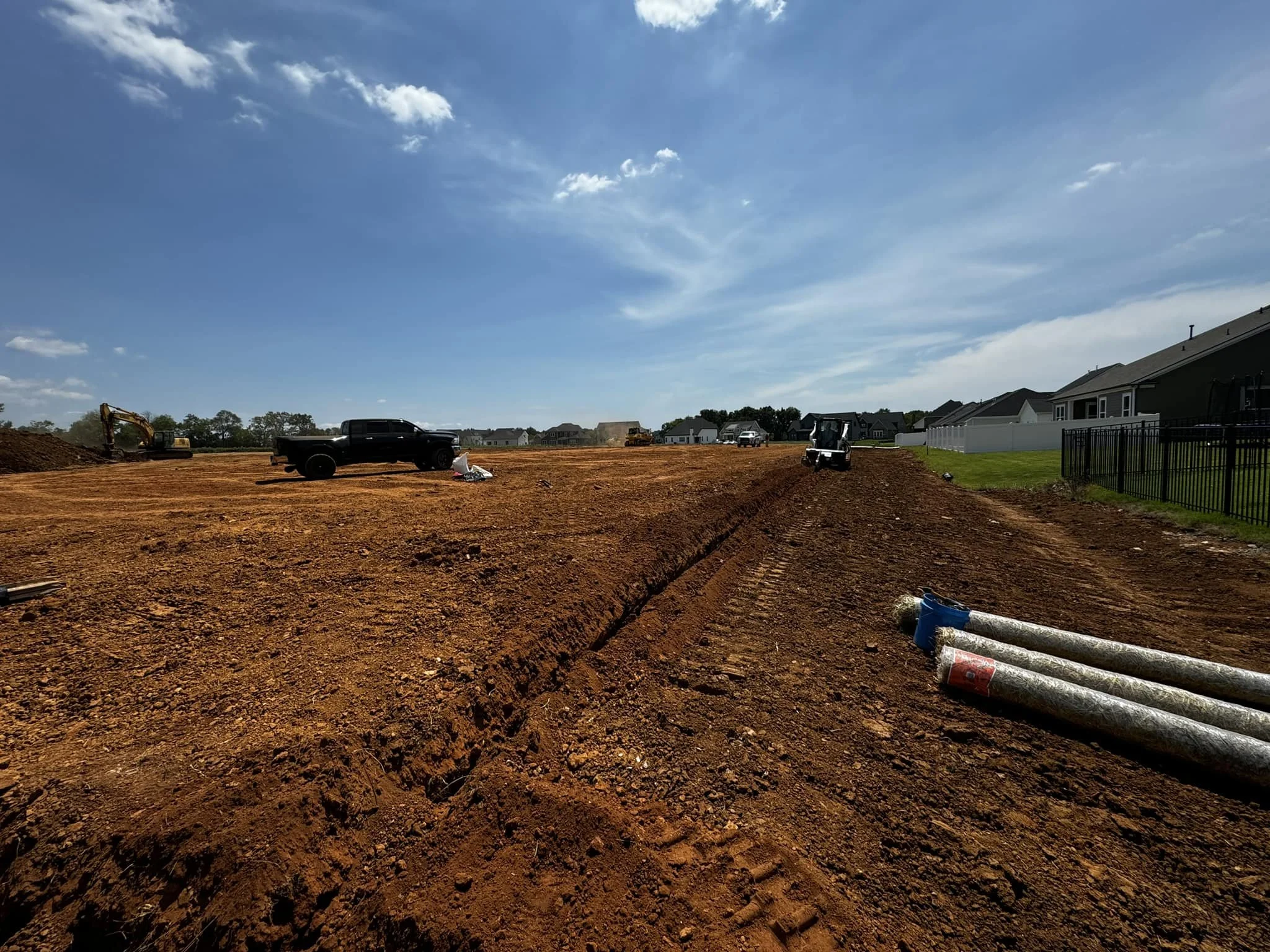 Construction site with dirt ground, construction vehicles, and residential houses in the background under a partly cloudy sky.