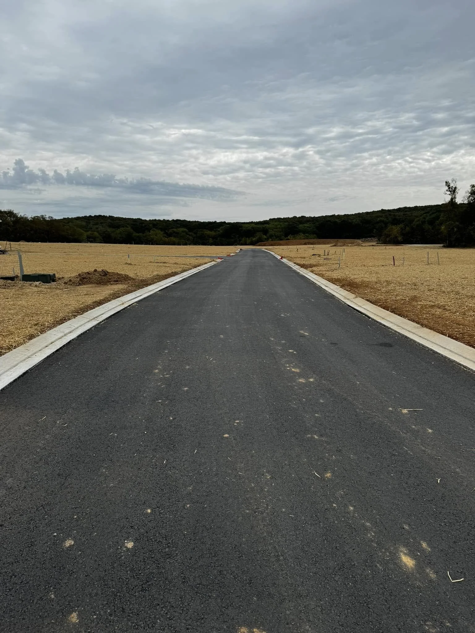 A newly paved asphalt road under a cloudy sky, leading into a rural landscape with trees and a hill in the distance, with dirt and construction materials on the sides.