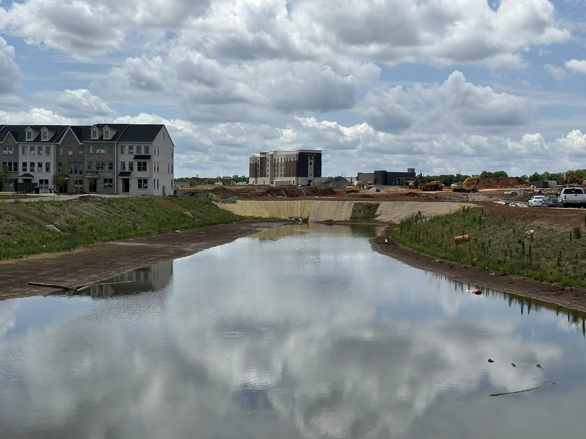 Construction site with buildings, dirt, and construction equipment next to a canal or small river, with cloudy sky overhead.