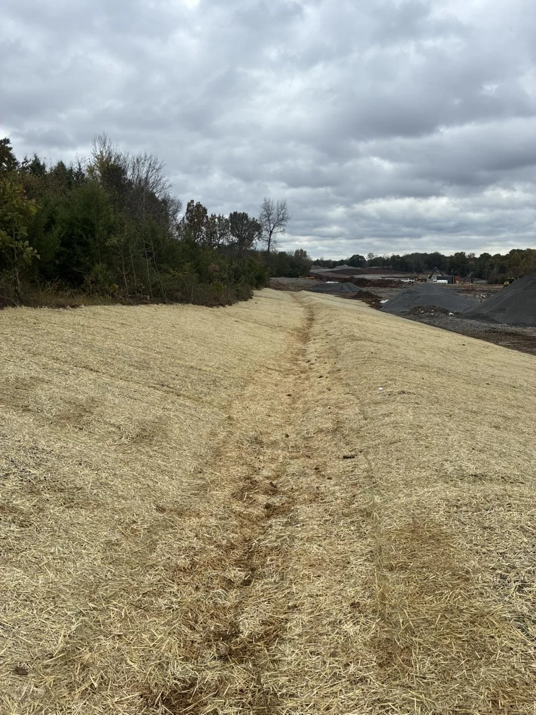 A dirt path runs through a large, open field with dry grass under a cloudy sky, with trees on the left and construction sites with piles of dirt on the right.