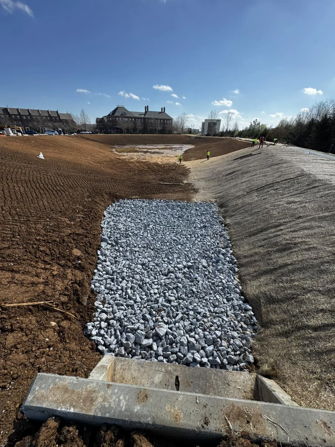 Construction site with gravel and dirt, workers visible in the background, and a building with a mansard roof and chimneys in the distance under a blue sky.