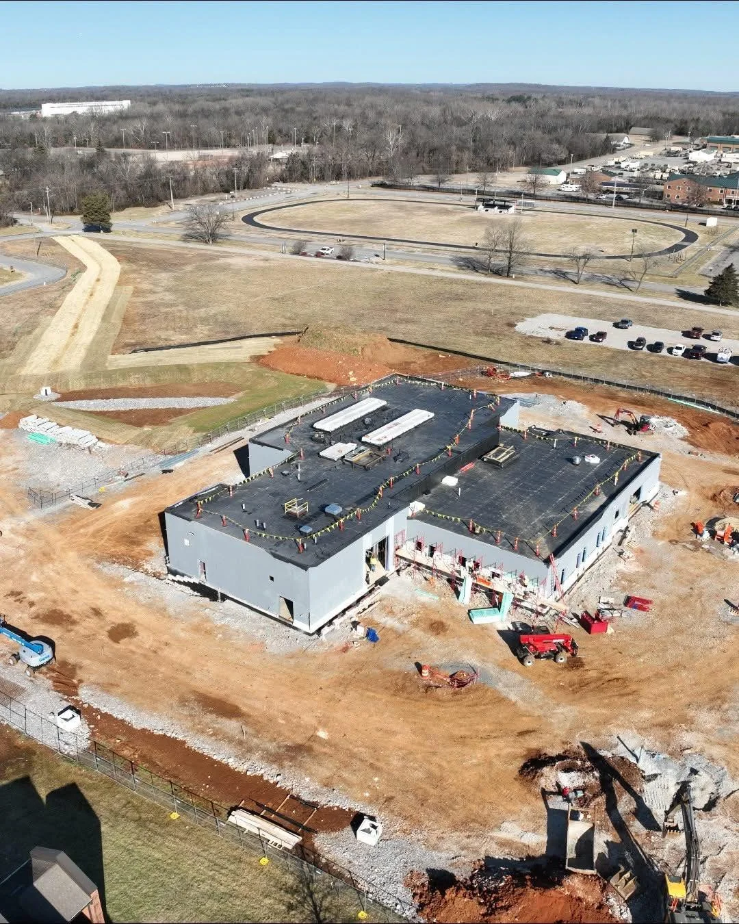 Construction site with a large building under construction, surrounded by dirt and construction equipment, with vehicles parked nearby and an athletic track in the background.