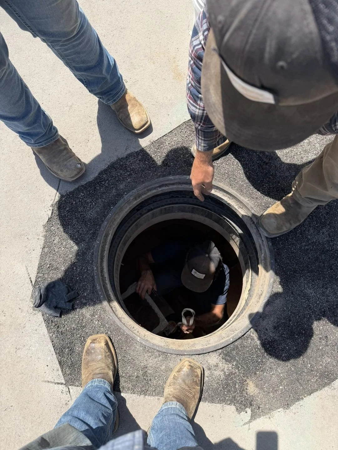 Four workers with cowboy boots and jeans or work clothes are gathered around an open manhole on a street, inspecting or working inside it.