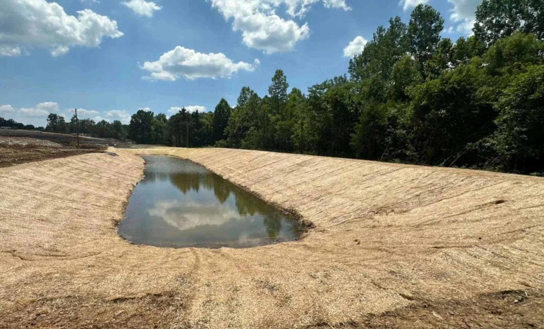 A small pond or water retention area with a shallow depth, surrounded by sloped, compacted sandy soil, with a line of trees and a partly cloudy sky in the background.