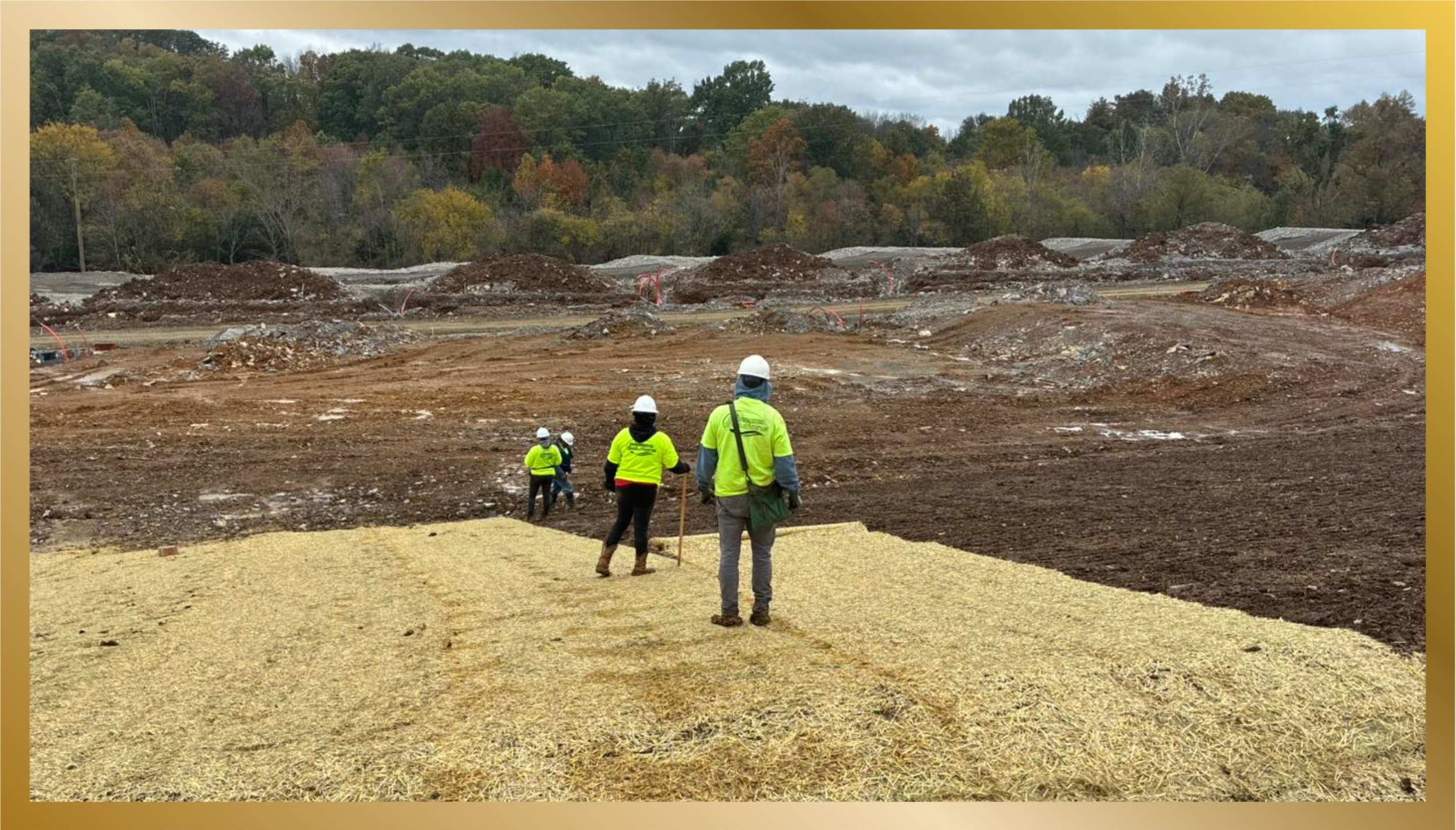 Construction workers in yellow safety vests and white helmets walking on a dirt-covered construction site with a forested area in the background.