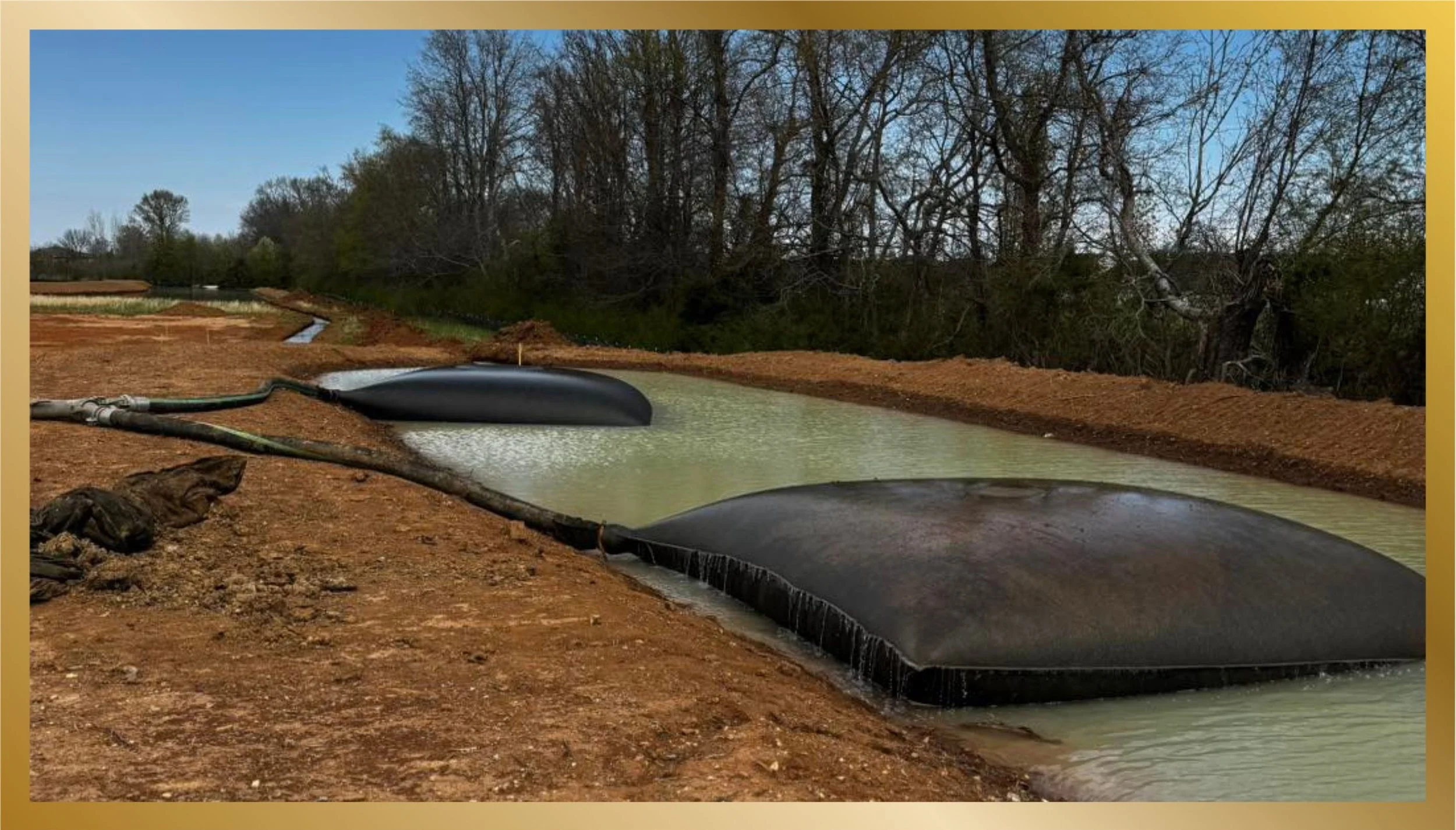 Construction site with large black water retention or filtration ponds filled with water, surrounded by dirt and pipes, with trees in the background.