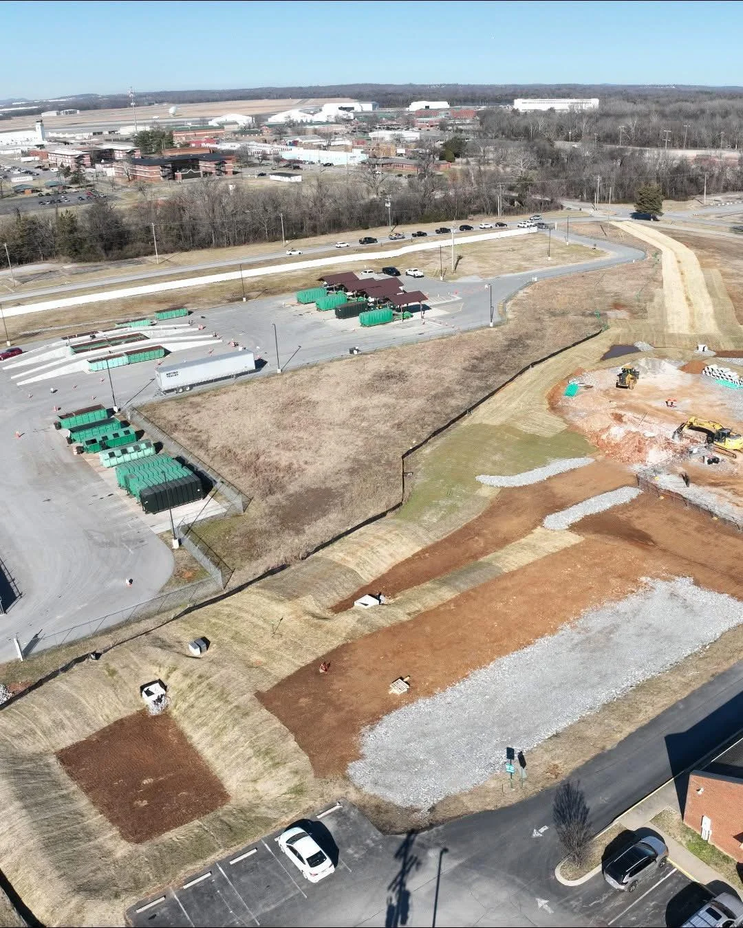 Aerial view of a construction site with excavation and groundwork, alongside parking lots, some green waste bins, and industrial buildings in the background.