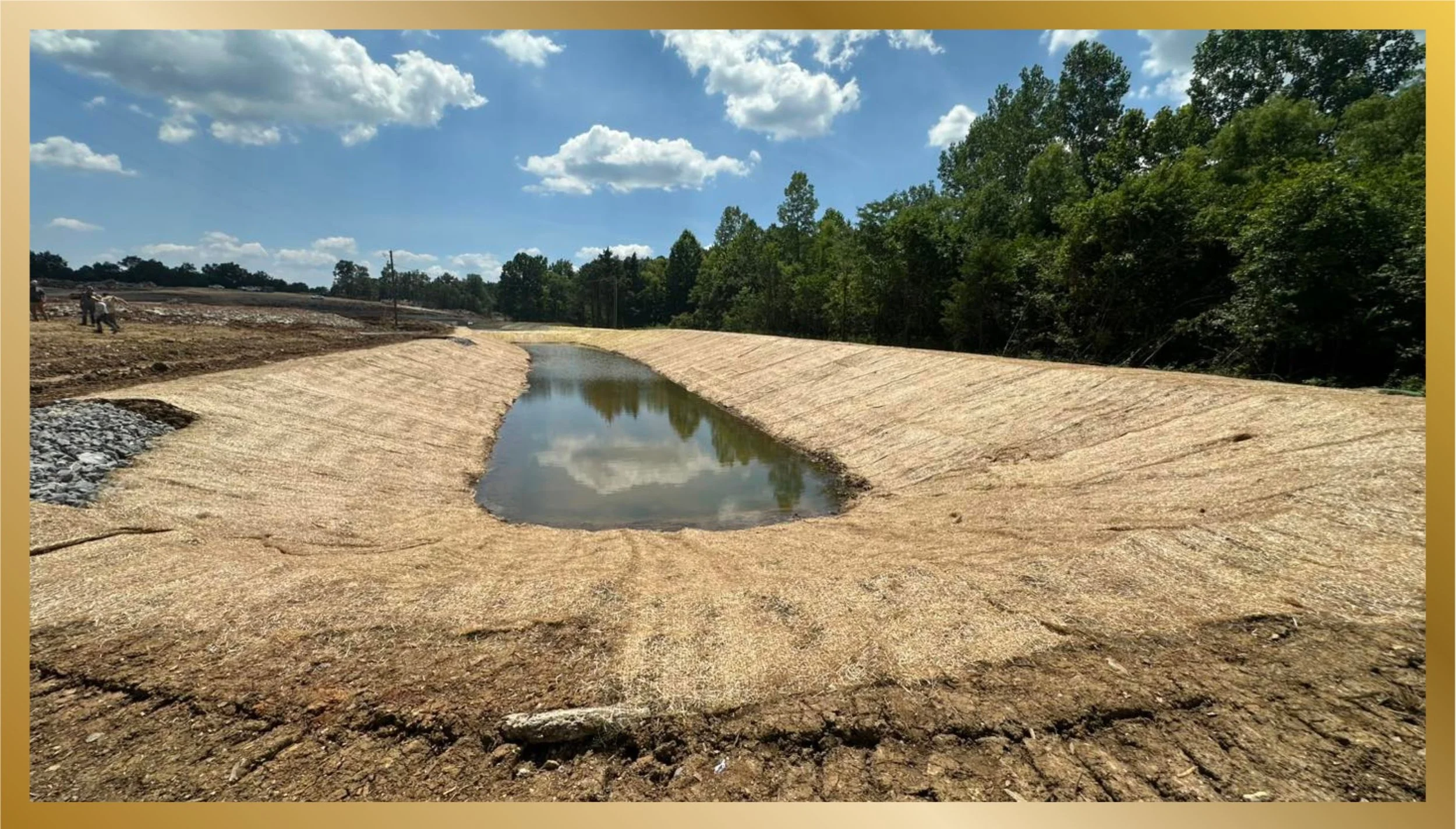 Construction site with a freshly paved canal or drainage ditch, reflecting the sky, surrounded by dirt and gravel with trees in the background.