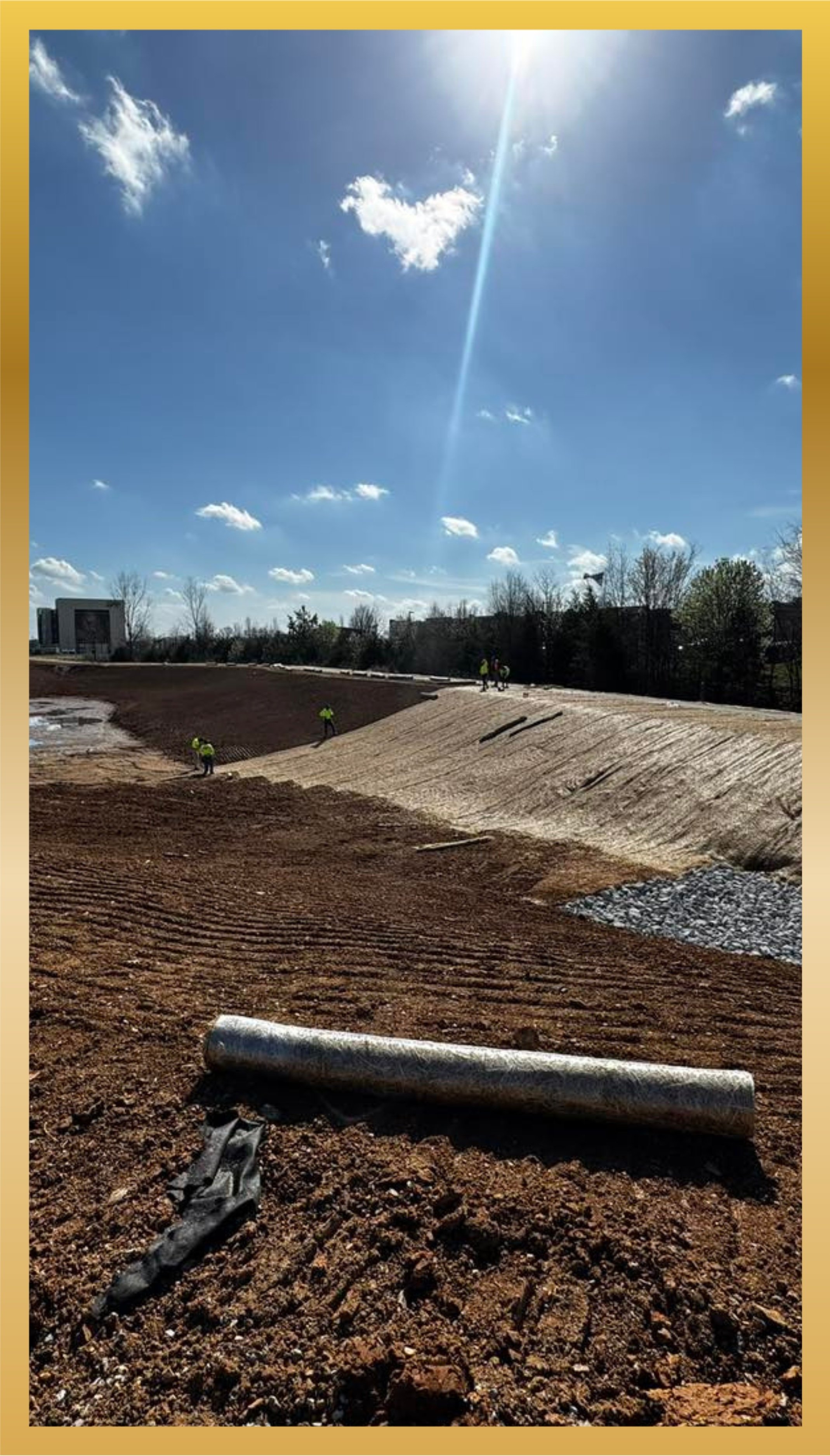 Construction site with workers on a dirt slope, pipe and debris in foreground, blue sky with clouds and bright sunlight overhead.