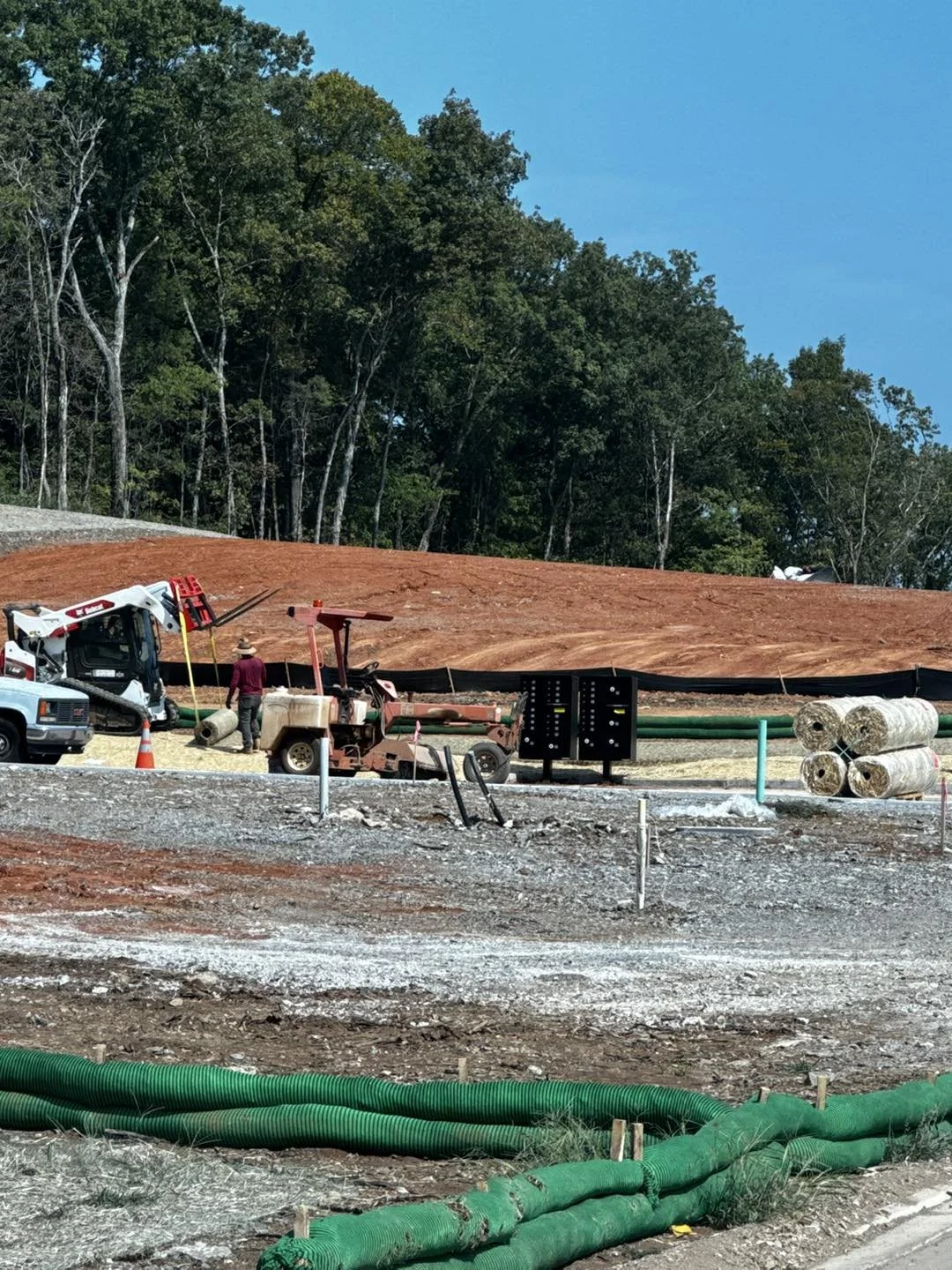 Construction site with machinery and rolled hay bales, large area of exposed soil, trees in the background, and a blue sky.