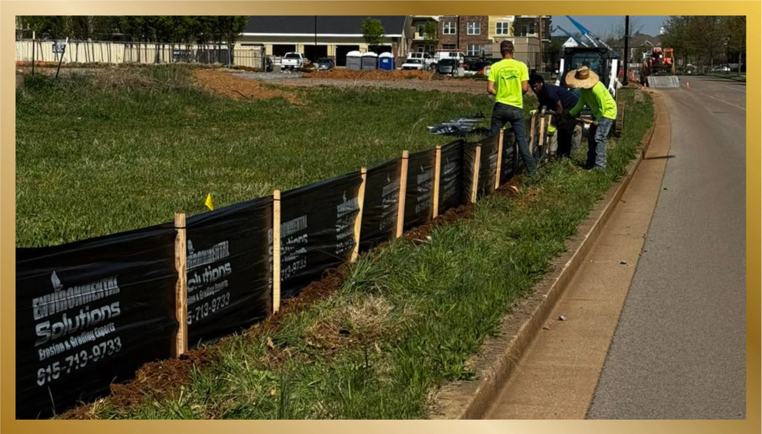 Construction workers installing a black landscape fabric along the edge of a grassy area, bordered by a sidewalk and a street, with machinery and parked cars in the background.