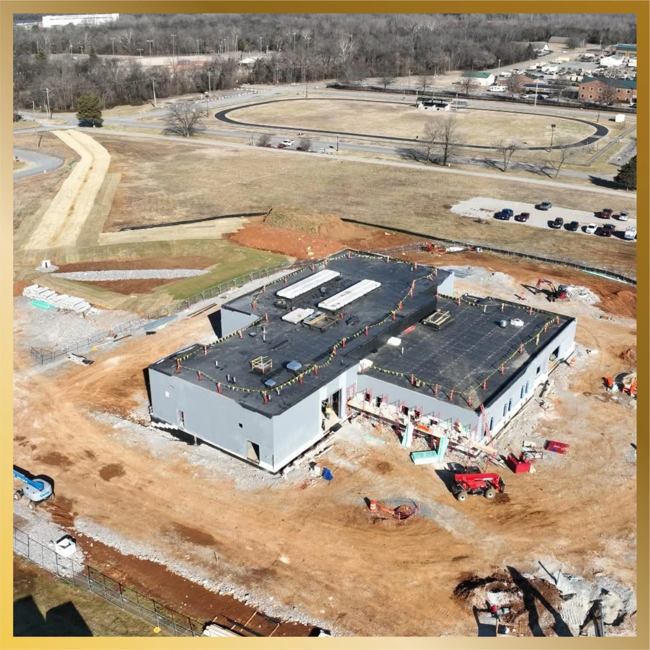 Aerial view of a construction site with a building in progress, parking lots, and landscaped areas, surrounded by trees and a wooded area in the background.
