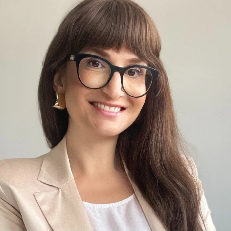 Eine lächelnde Frau mit braunen Haaren, Brille und eleganter Kleidung.