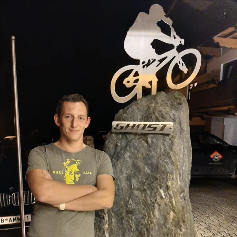 Ein Mann steht vor einem großen Felsen mit einem Metalllogo, das den Markenname „GHOST“ trägt, und einer metallenen Fahrrad-Silhouette im Hintergrund bei Nacht.