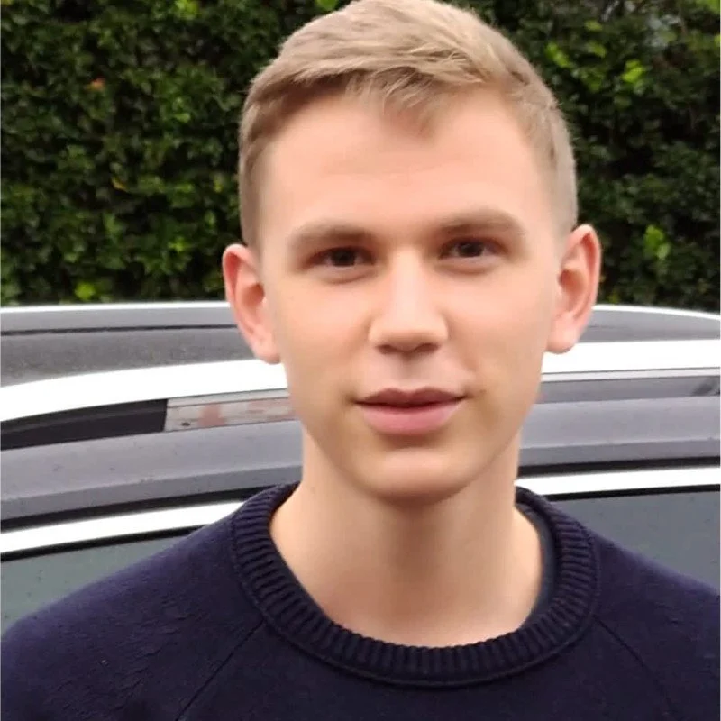Junge Mann steht vor einem schwarzen Auto mit einem grünen, bewachsenen Hintergrund.