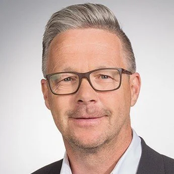 Porträt eines Mannes mit Brille, grauem Haar und gepflegtem Bart vor hellem Hintergrund.