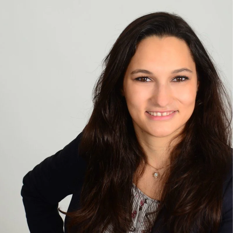 Junge Frau mit langen dunklen Haaren, lächelnd, im professionellen schwarzen Blazer vor hellem Hintergrund.