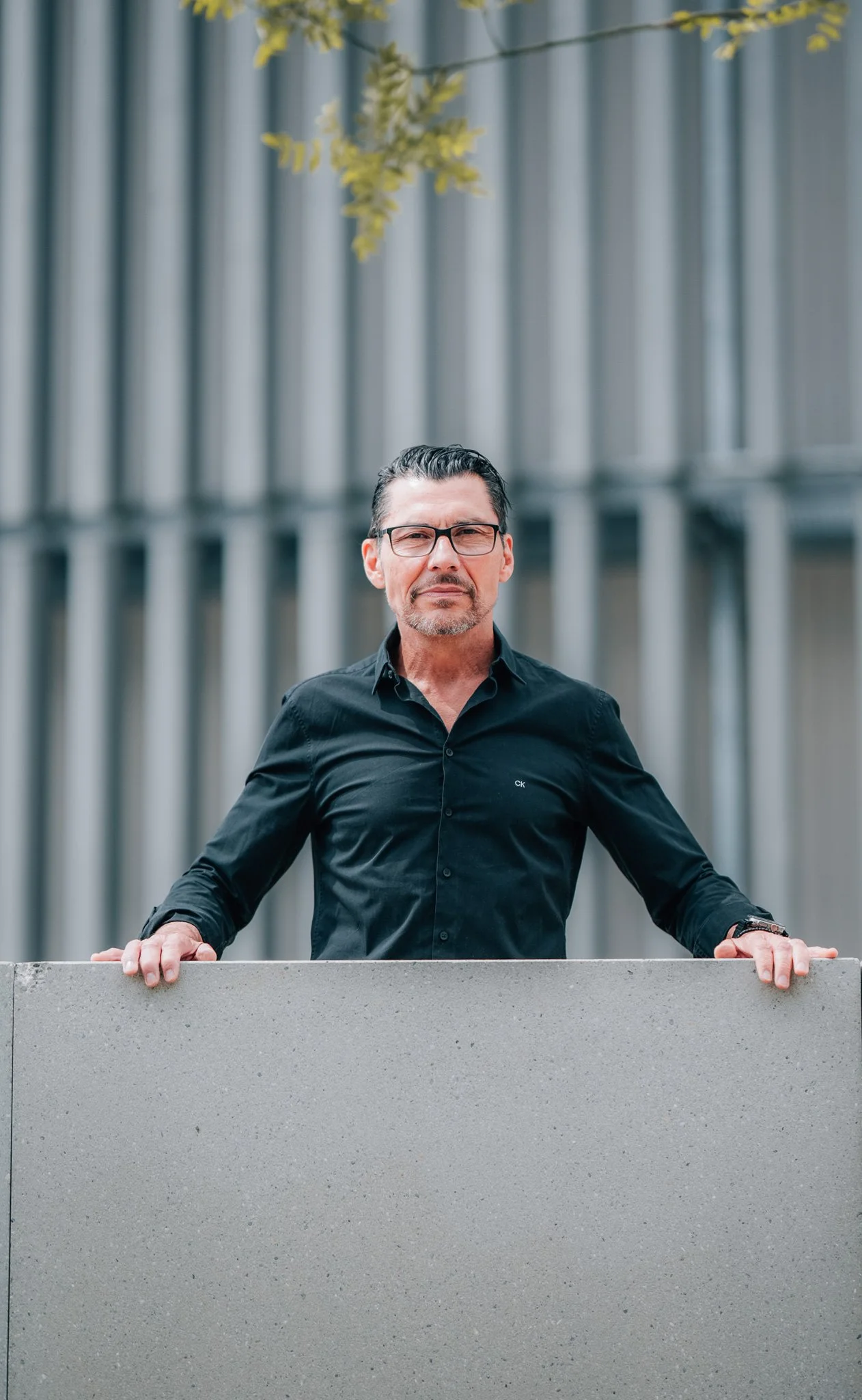 Ein Mann mit schwarzen Haaren, Brille und Bart steht vor einer modernen Gebäudefassade aus Metall und hält sich mit beiden Händen an einer niedrigen Betoneinfassung fest.