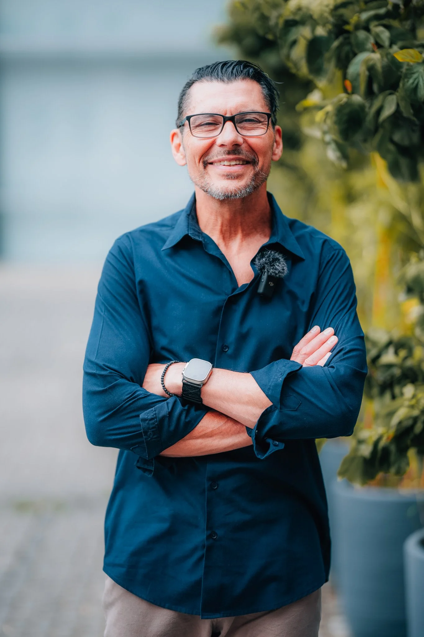 Ein lächelnder Mann mit dunklen Haaren, Brille und grauem Bart, trägt eine dunkelblaue Hemd, steht vor grünen Pflanzen, mit einem weißen Smartwatch am linken Handgelenk, auf einem Weg im Freien.