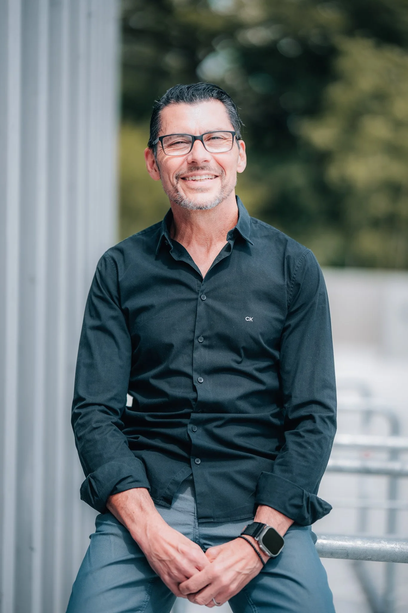 Ein lächelnder Mann mit schwarzen Haaren, Brille, dunklem Hemd und Armbanduhr sitzt auf einer Bank im Freien, im Hintergrund sind Bäume und ein Gebäude sichtbar.