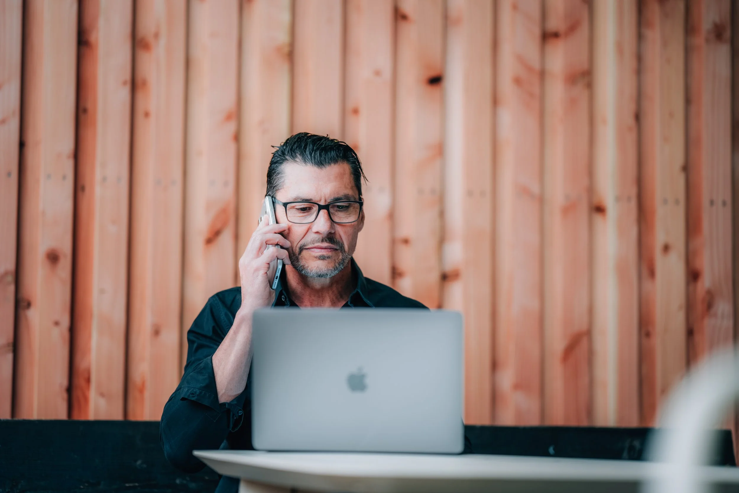 Mann mit Brille telefoniert während er an einem Laptop arbeitet, im Hintergrund Holzvertäfelung.