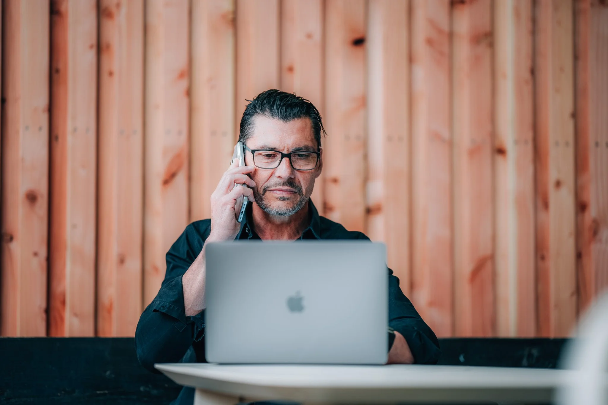 Mann mit Brille beim Telefonieren vor einem Laptop, im Hintergrund eine Holzvertäfelung.