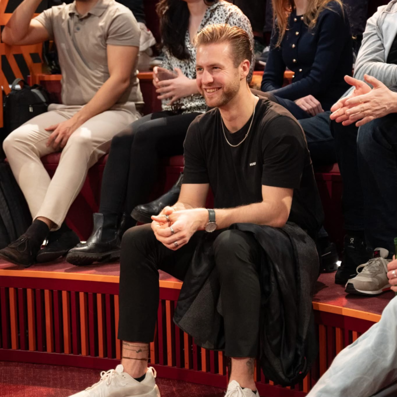 Ein junger Mann mit kurzer blonder Haar, schwarzem T-Shirt, schwarzen Hosen und weißen Sneakern sitzt lächelnd im Publikum bei einer Veranstaltung.