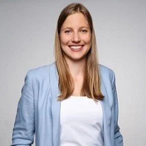 Ein Lächeln, eine Frau mit langem braunem Haar, blauer Blazer und weißem Top vor grauem Hintergrund.