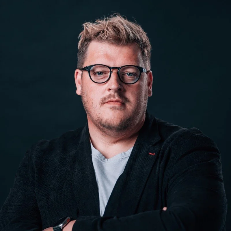 Porträt eines Mannes mit Brille, grauer T-Shirt, schwarzem Jacke vor dunklem Hintergrund.