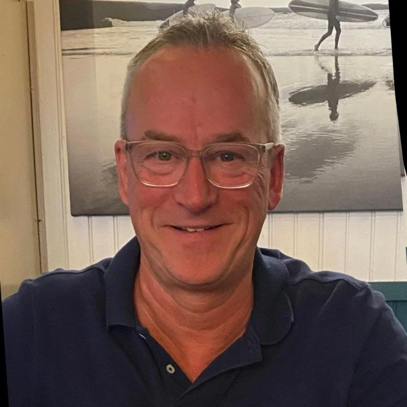 Ein lächelnder Mann mit Brille in einem dunklen Polo-Hemd sitzt in einem Raum mit einer Wand, auf der ein Schwarz-Weiß-Bild von Surfern am Strand hängt.