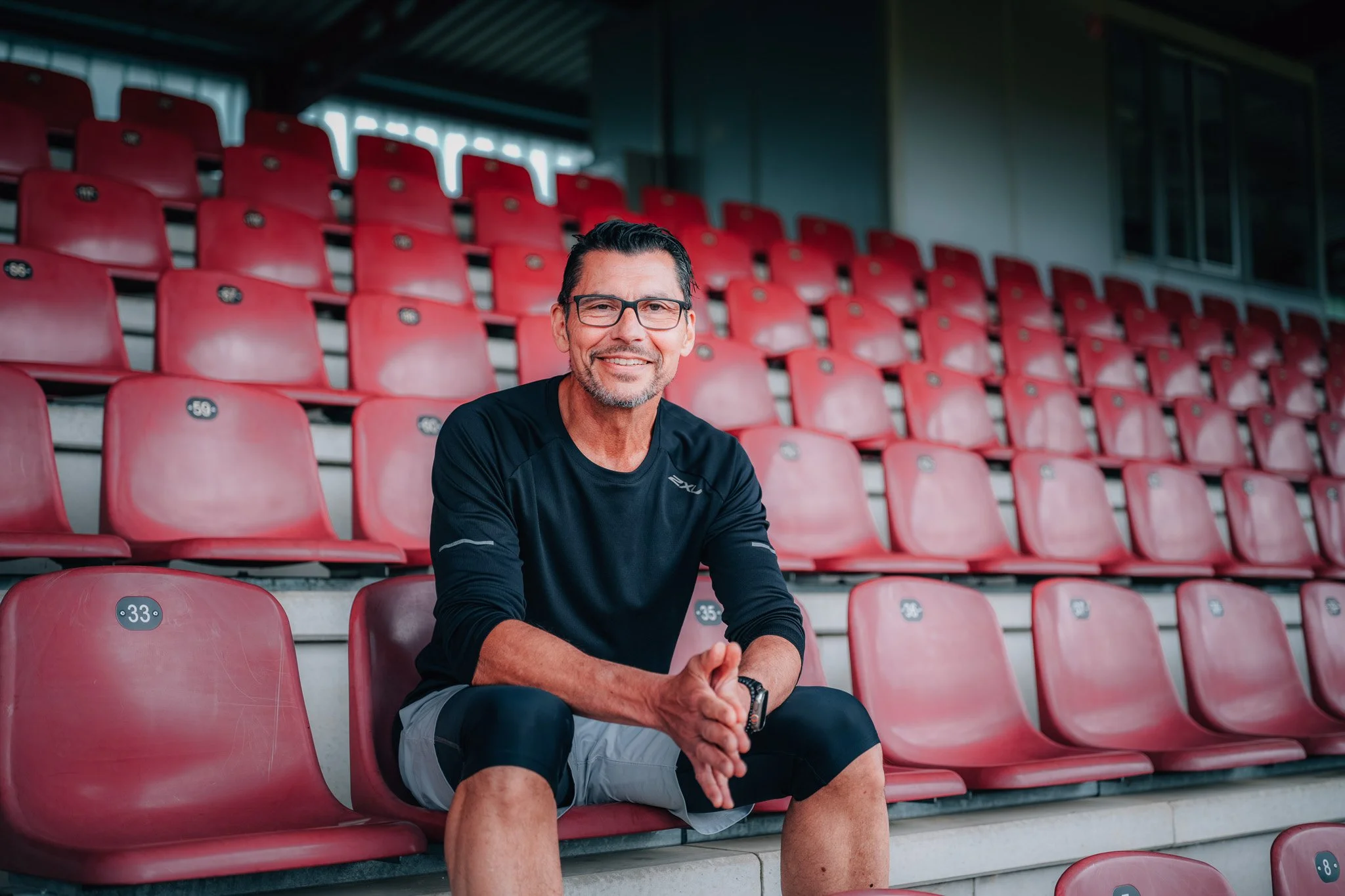 Ein lächelnder Mann mit Brille sitzt auf roten Stadionbänken in einem leeren Stadion, trägt ein schwarzes Sportshirt und graue Shorts.