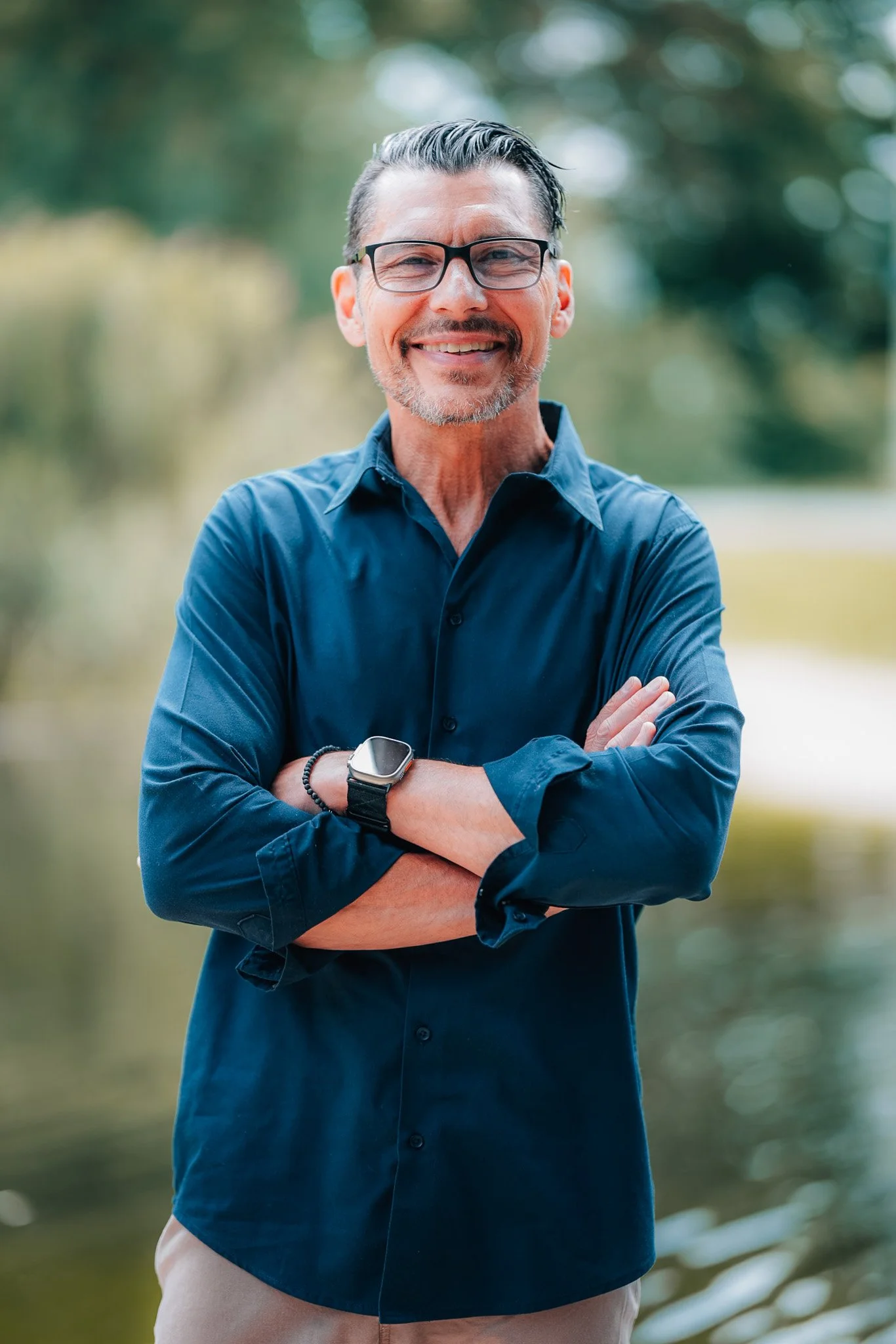 Ein lachender Mann mit Brille steht im Freien vor einem Wasser im Hintergrund, trägt eine dunkelblaue Hemd und eine Smartwatch am Handgelenk.