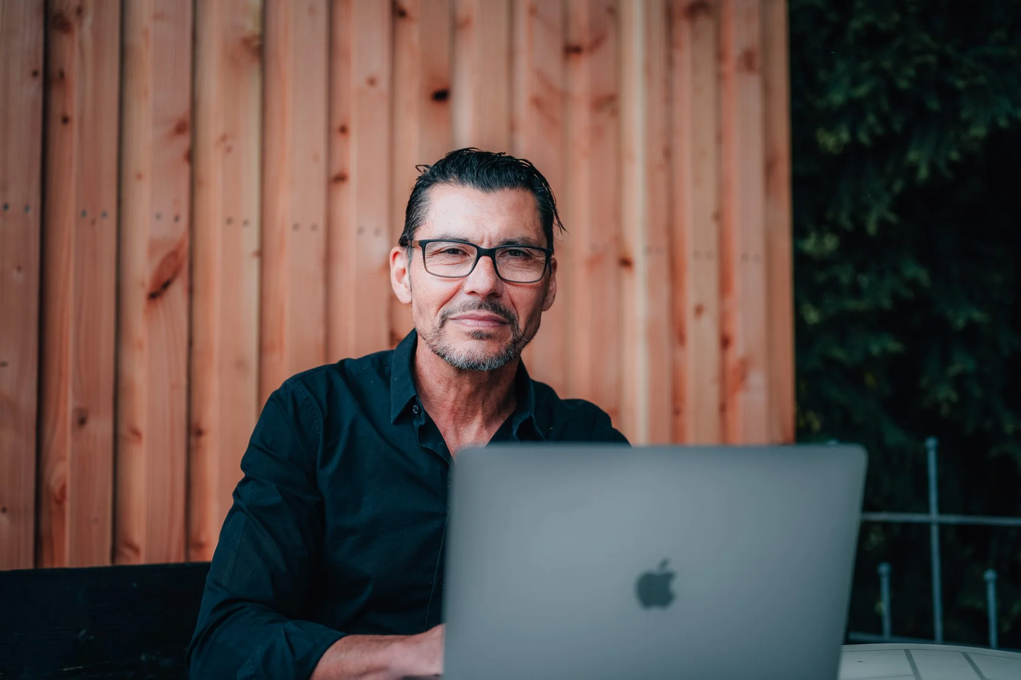 Ein Mann mit bräunlich-schwarzem Haar und Brille sitzt vor einem Laptop und schaut in die Kamera, im Hintergrund eine Holzwand und Bäume.