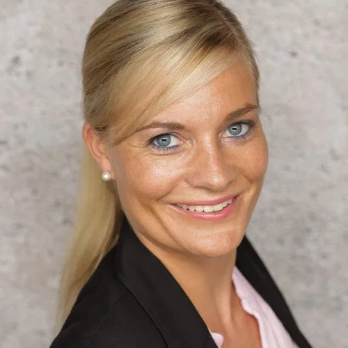Porträt einer Frau mit blonden Haaren, blauer Augen, pearllohrringen und schwarzer Jacke, vor einer grauen Wand.