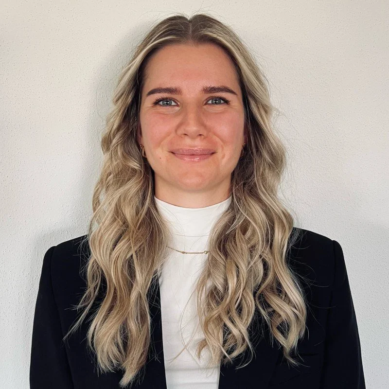 Ein Lächelndes junges Frau mit langen, lockigen, blonden Haaren, in professioneller Kleidung, vor einer weißen Wand.