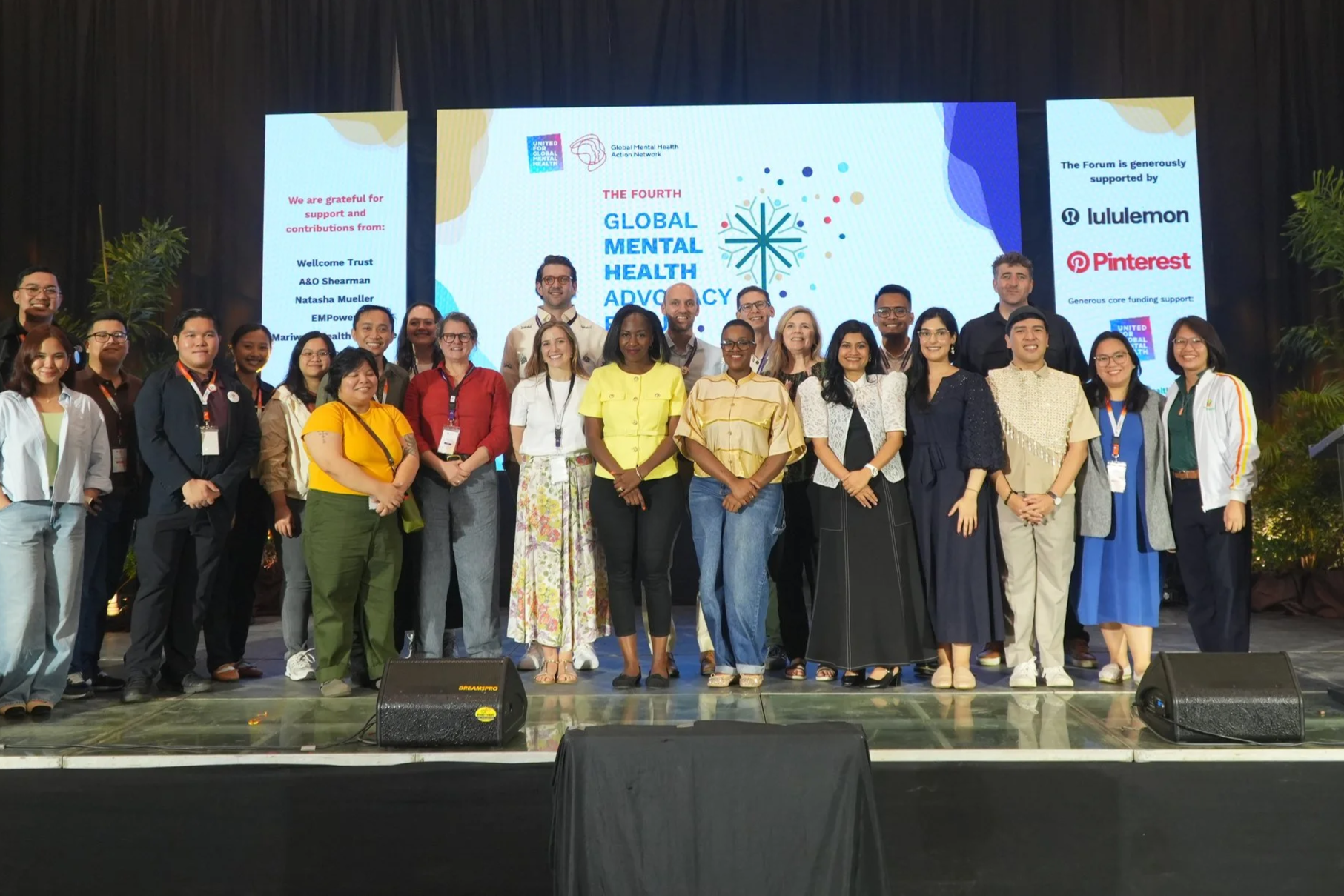 A group of diverse people posing on stage at a conference or event. Behind them is a large screen displaying the text 'The Fourth Global Mental Health Advocacy Forum' along with logos and supporting sponsor names like Lululemon and Pinterest.