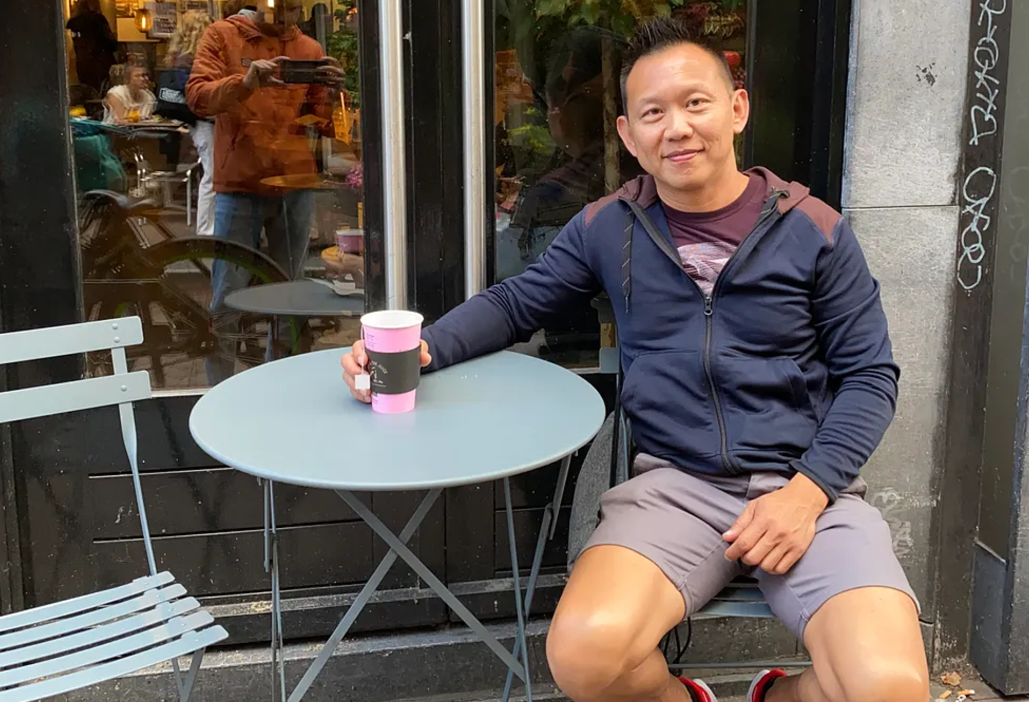 A man sitting outside at a cafe, holding a pink to-go coffee cup, smiling, wearing a navy blue jacket, gray shorts, and red sneakers. The cafe has a black window with graffiti on the side, and there is a reflection of a woman taking the photo in the window.