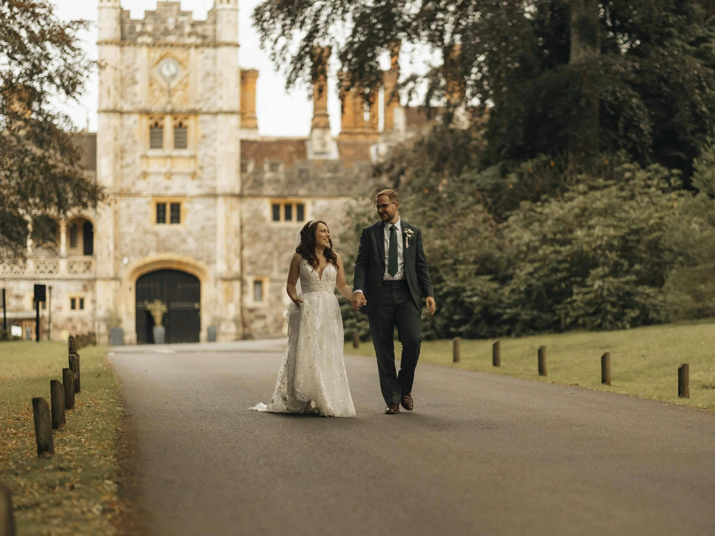 A bride and groom holding hands and walking on a paved path in a park, with a historic castle in the background and trees surrounding them.