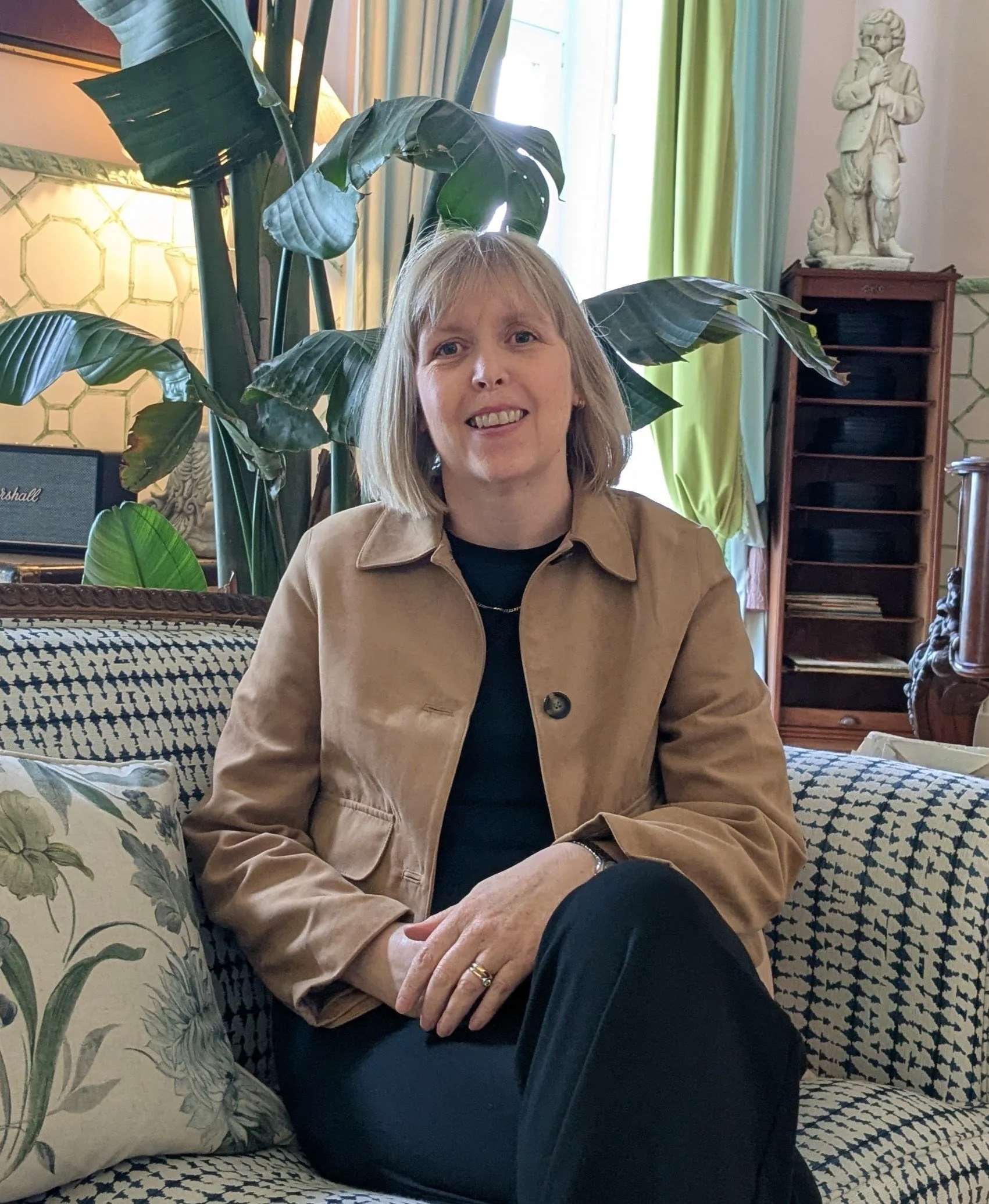 A woman with blonde hair sitting on a patterned couch in a room with green curtains, a large plant in the background, and statues on a shelf. She is wearing a tan jacket and black pants.