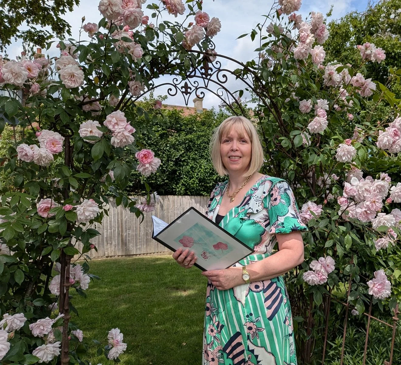 Woman standing in a garden with pink roses, holding a folder, wearing a colorful patterned dress and gold jewelry.