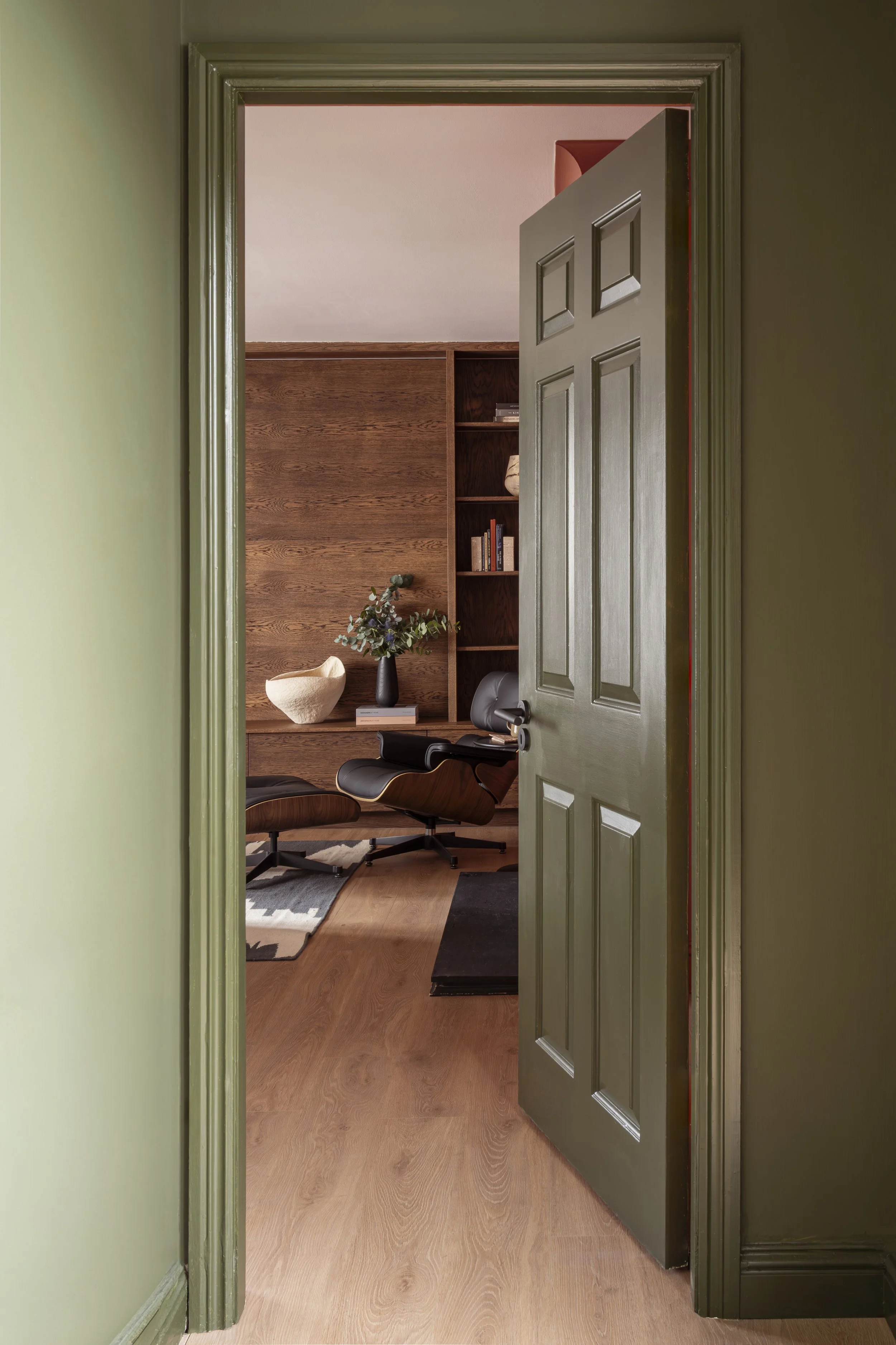 View through the hallway of the Portobello, a partially open green door into the living area with wood flooring, bespoke dark wood joinery unit, , a modern lounge Eames armchair. Joinery unit styled with a vase with flowers and other ceramics.