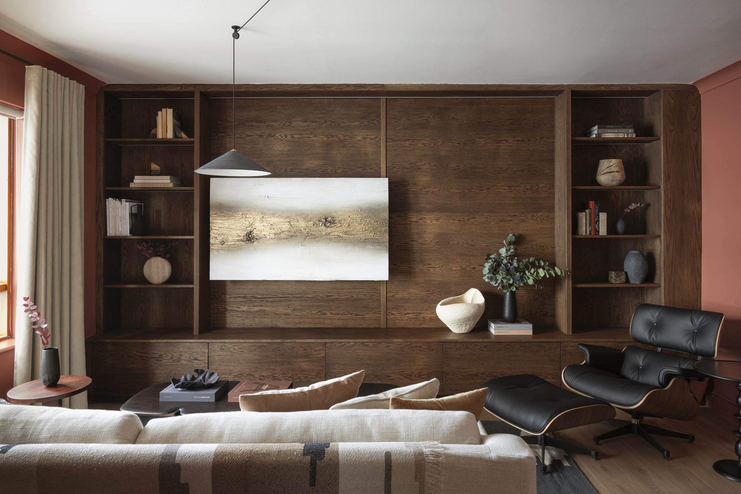 Living room with bespoke wood joinery unit, oatmeal fabric sofa dressed with a patterned throw and cushions, sculptural black leather Eames armchair, abstract artwork and a coffee table styled with books — interior design by Susan Cotter Design.