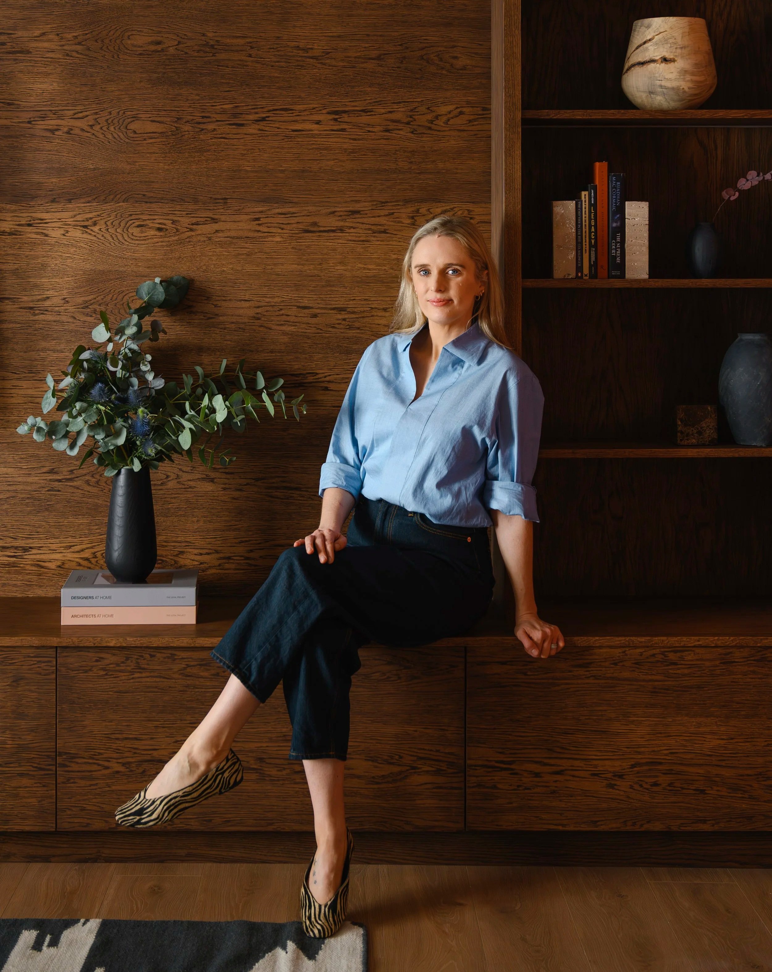 Susan Cotter, founder of Susan Cotter Design, seated on the bench of a bespoke joinery unit with built-in drawers and open shelves. Open shelves styled with ceramics and books.
