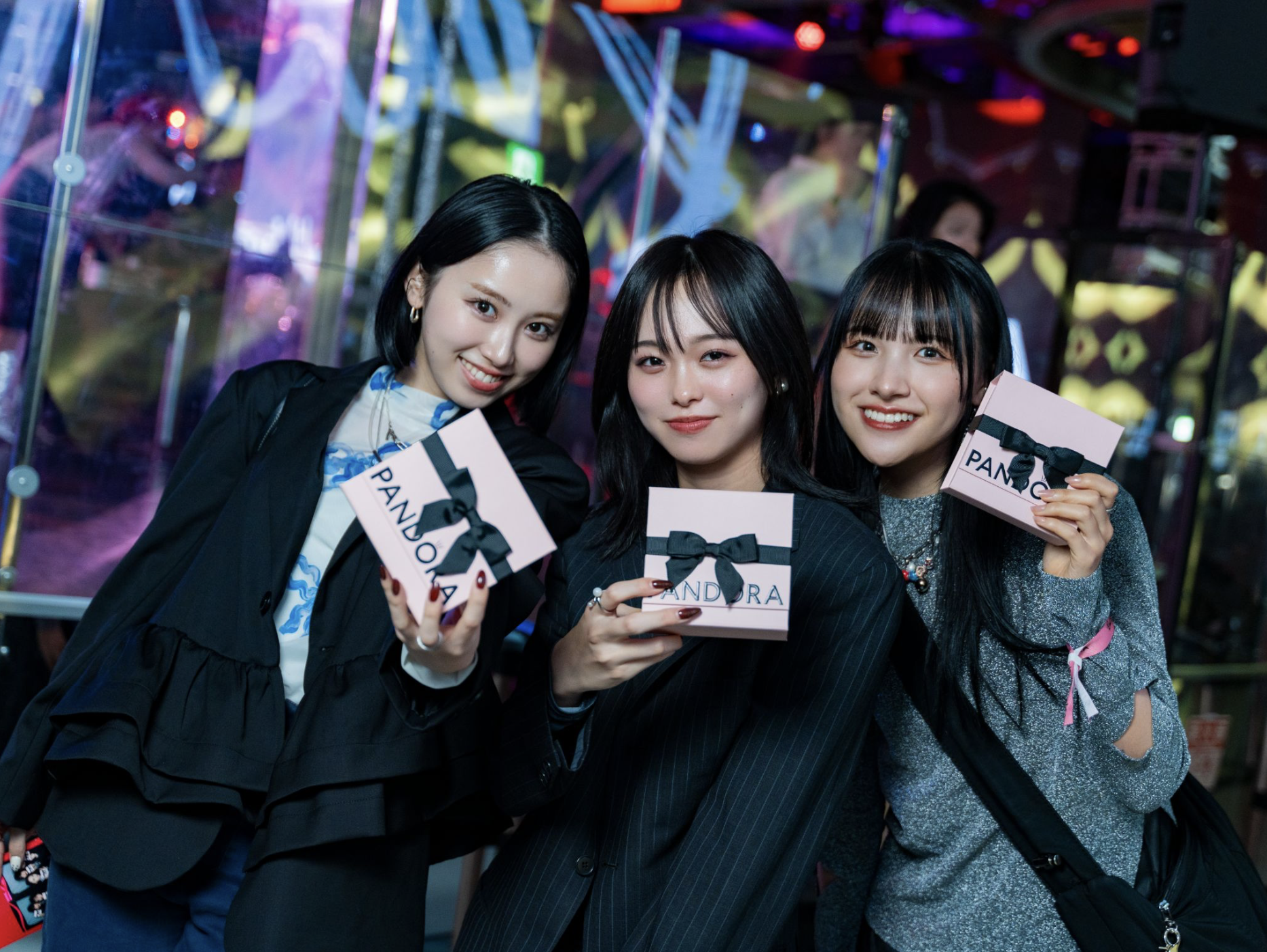 Three young women smiling and holding pink gift boxes with black ribbons in a colorful, lively indoor setting with vibrant lights and reflections.