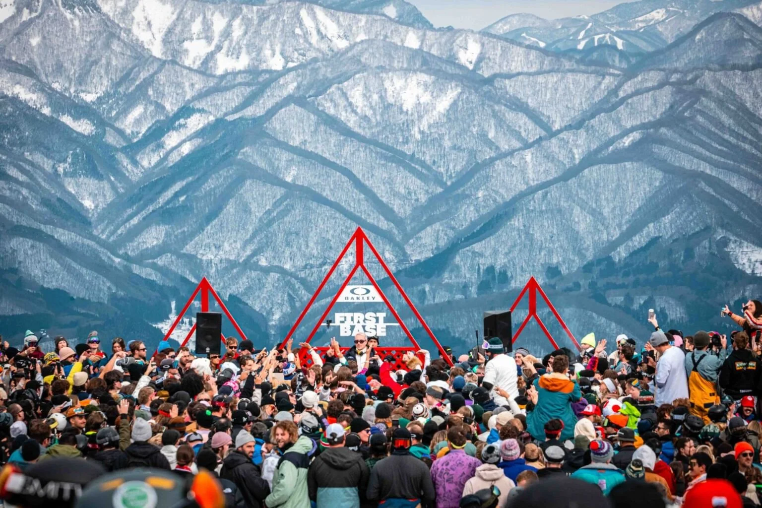 Large crowd gathered outdoors at a concert or event with a stage, mountains in the background, and many people taking photos or videos.