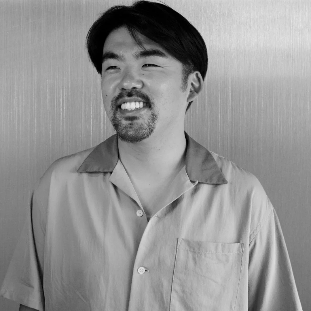 A smiling man with a beard and mustache, wearing a collared shirt, standing against a plain background in black and white.
