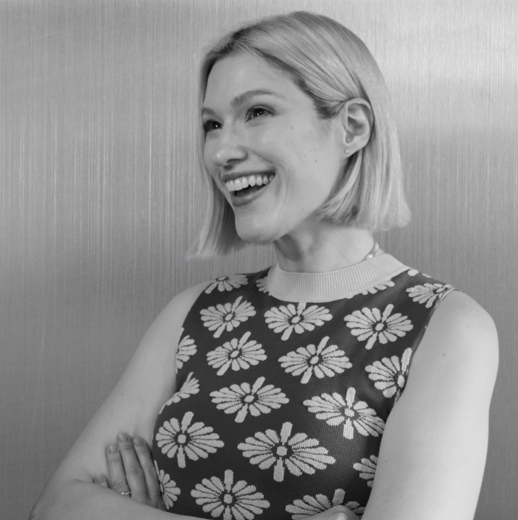 A woman with short blonde hair smiling and looking to the side, wearing a sleeveless dress with a daisy flower pattern, standing in front of a plain wall.